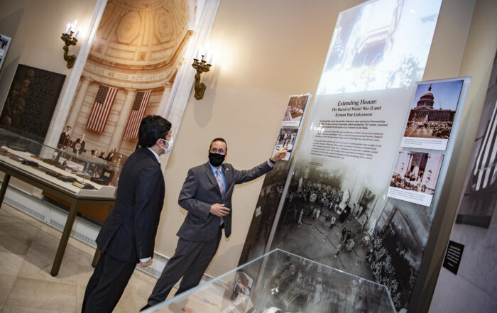 Tim Frank (right), historian, giving a tour of the Memorial Amphitheater Display Room exhibit to State Minister of Defense of Japan Nakayama Yasuhide (left) on August 10, 2021. One of the large glass panels with text, images, and original motion picture footage being projected on to it can be seen.