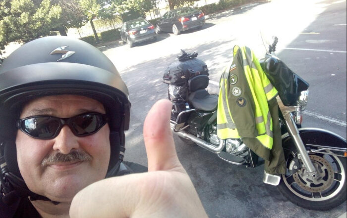 John Sterkendries wears a helmet and sunglasses and does a thumbs-up next to his motorcycle