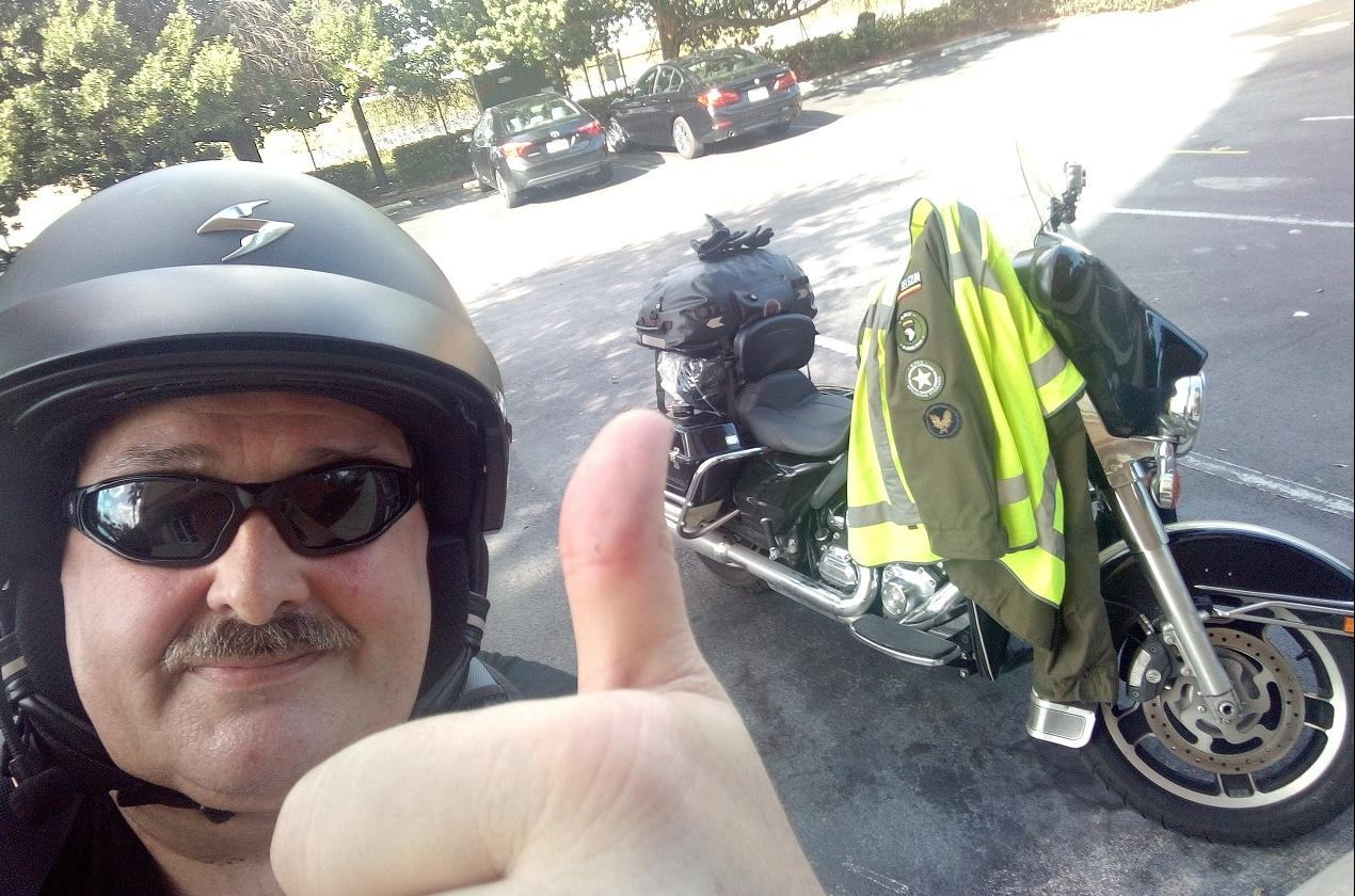 John Sterkendries wears a helmet and sunglasses and does a thumbs-up next to his motorcycle