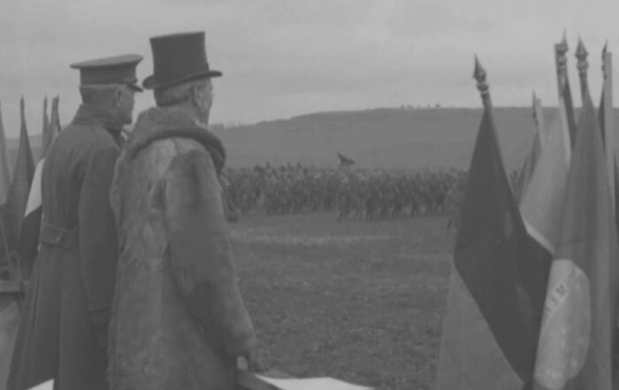 Two men in hats and coats review a group of soldiers in a field.