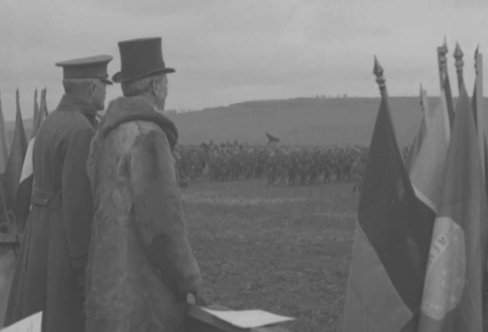 Two men in hats and coats review a group of soldiers in a field.