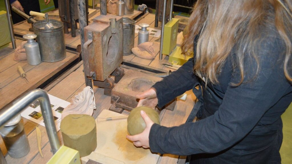 A blond person in a black jacket makes a clay shape at a table