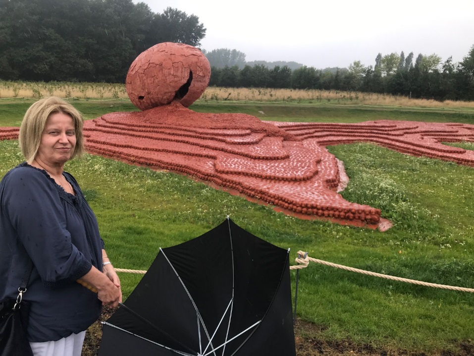 A blonde person with an umbrella stands near the clay figurine memorial