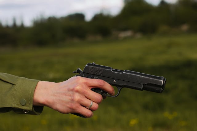 A hand of a man in a green shirt, holding an M1911 pistol with blurred greenery in the background.