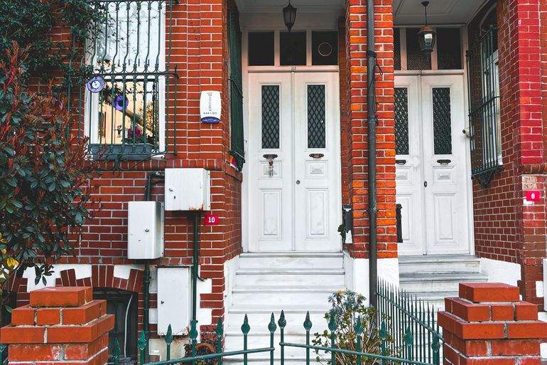 A red brick twin rowhouse facade with white double doors, iron window grilles, and potted plants, typical of older Philadelphia residential blocks.