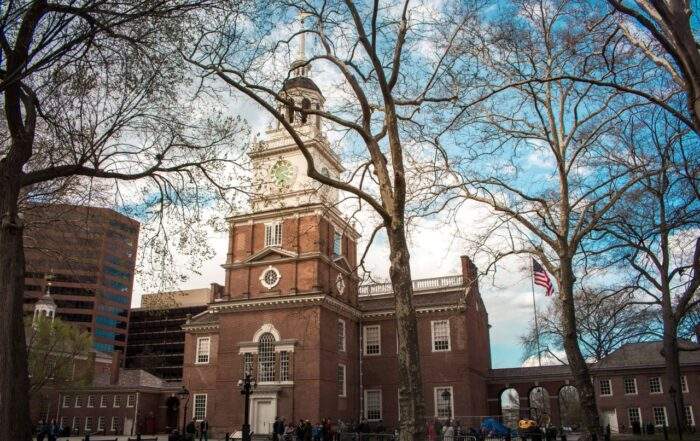 Independence Hall in Philadelphia, framed by bare trees in early spring, with an American flag visible to the right of the building.
