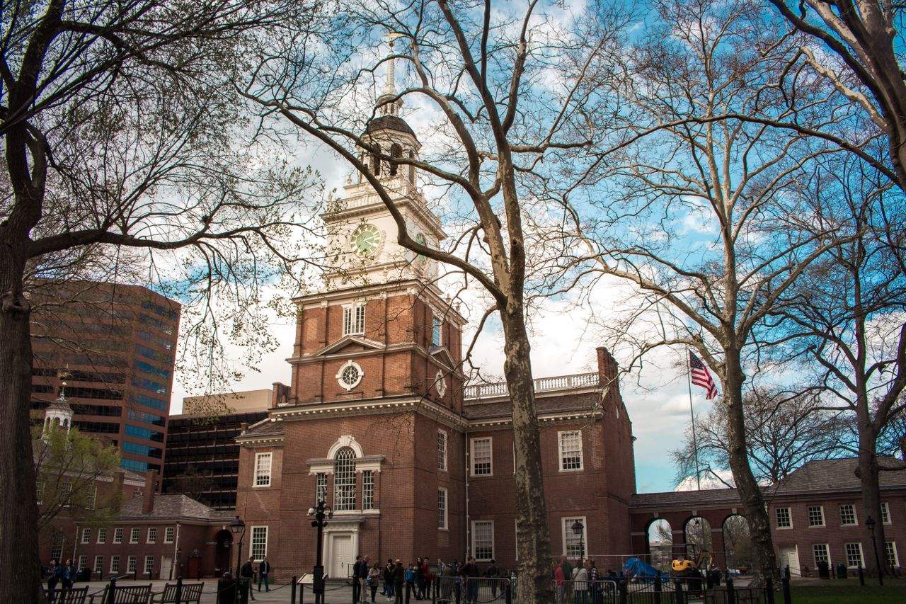 Independence Hall in Philadelphia, framed by bare trees in early spring, with an American flag visible to the right of the building.